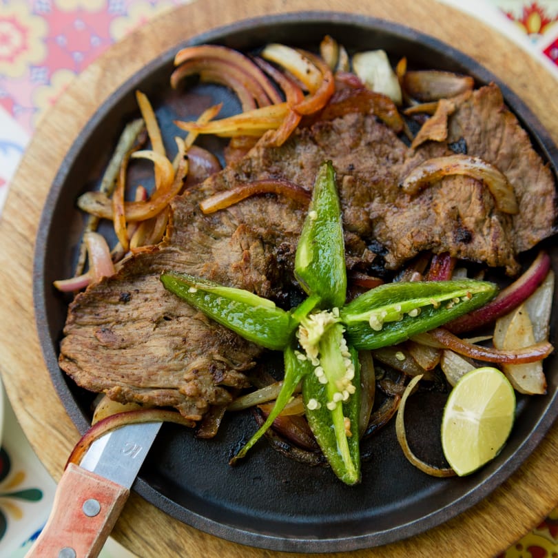 Sizzling steak fajitas with grilled onions, jalapeños, and a lime wedge in a cast iron skillet.