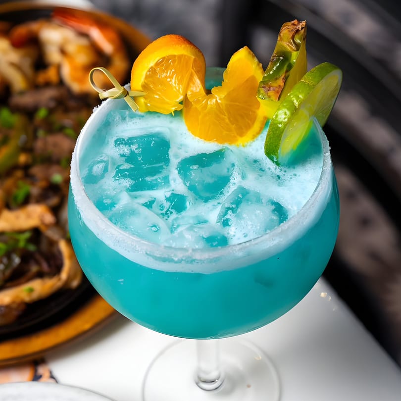 A vibrant blue El Paso Tacos & Tequila margarita with ice, garnished with orange, lime, and pineapple.