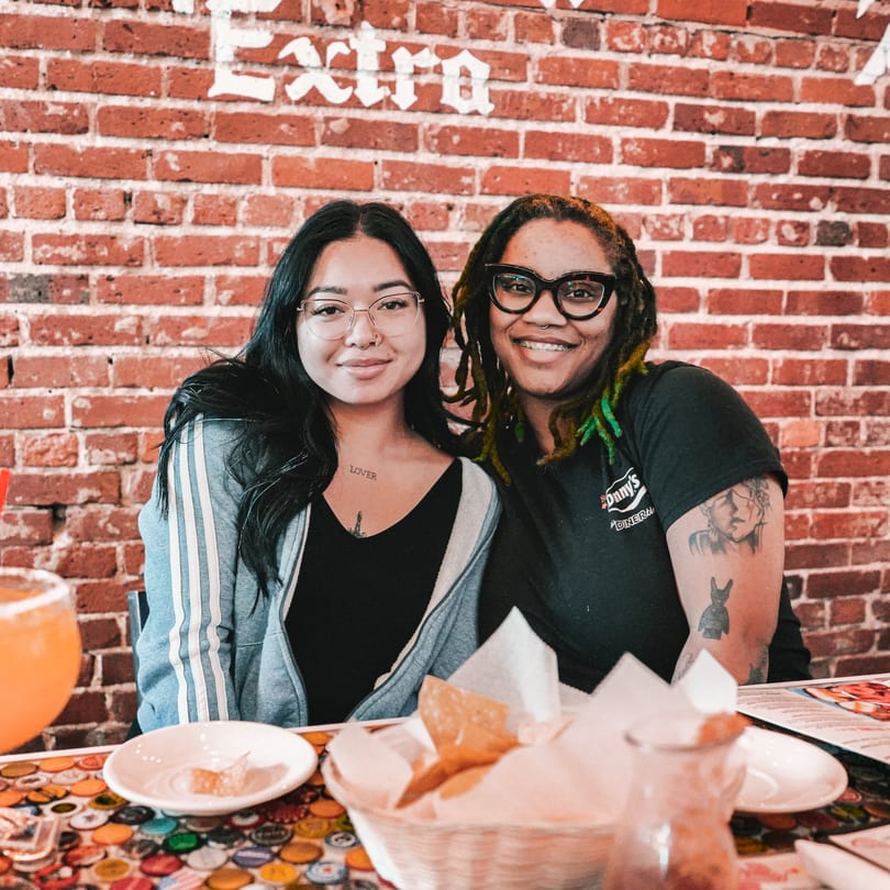 Two smiling women sit at a table with chips and drinks, against a brick wall.