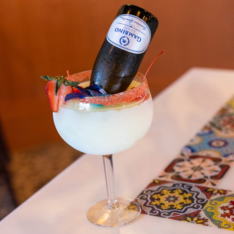 A large margarita with a mini Gambino Cuvee bottle, strawberry, and cherry on a white table.