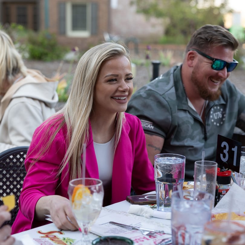 Smiling woman in a pink blazer at an outdoor table with drinks and a man in sunglasses.