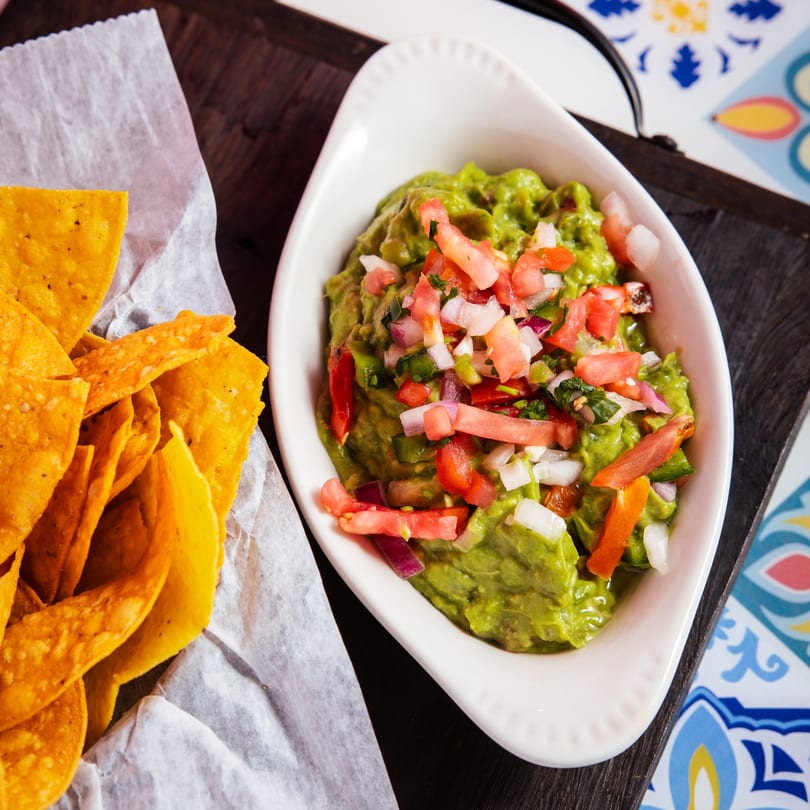 A white dish of guacamole topped with pico de gallo next to a basket of tortilla chips.