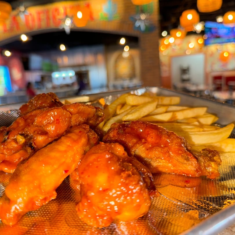 Spicy chicken wings and french fries on a metal tray, with a blurred restaurant interior in the background.