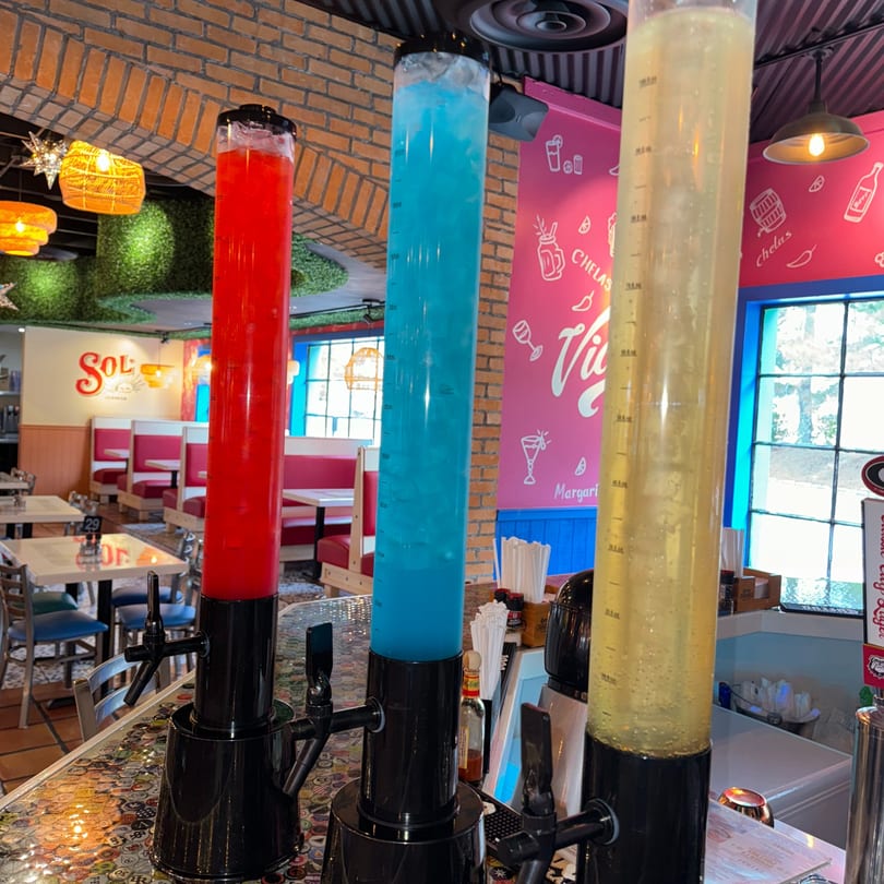 Three colorful margarita towers, red, blue, and yellow, on a bar in a vibrant Mexican restaurant.